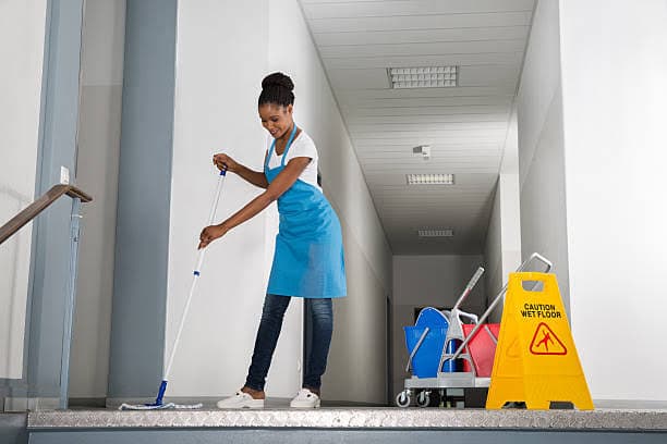 Smiling woman in a blue apron mopping a hallway floor near a wet floor sign.
