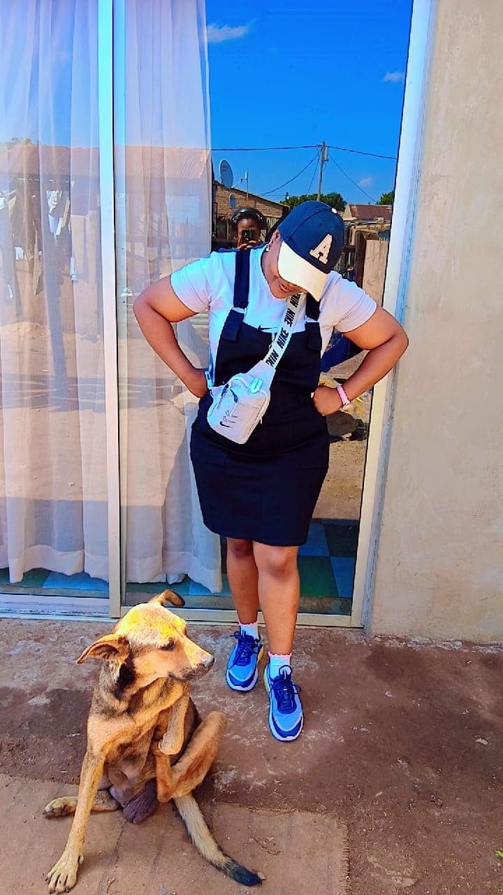 Woman in black overalls and blue cap posing beside a sitting brown dog outdoors.