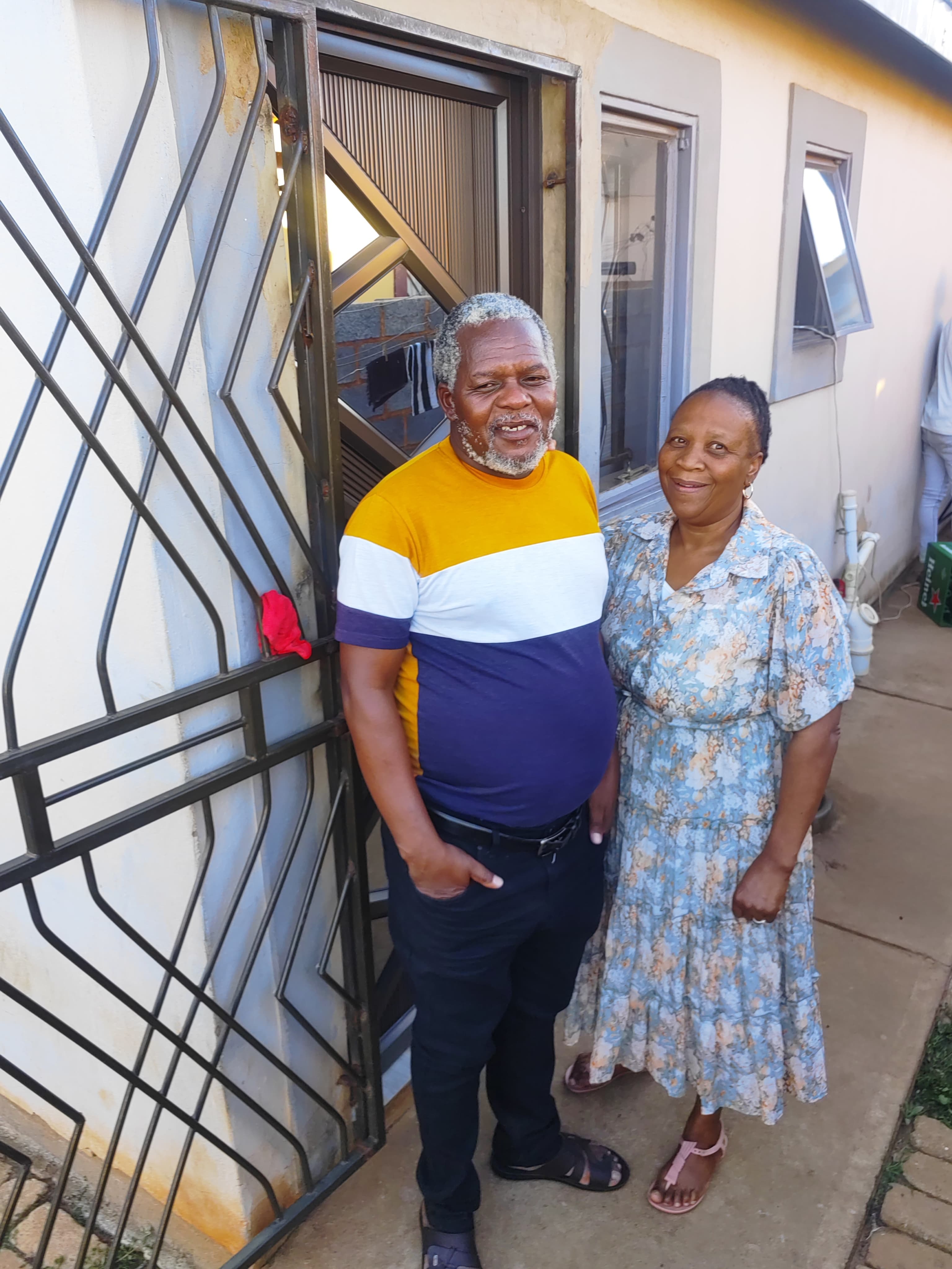 Smiling older couple standing together outside a house next to a black metal security gate.