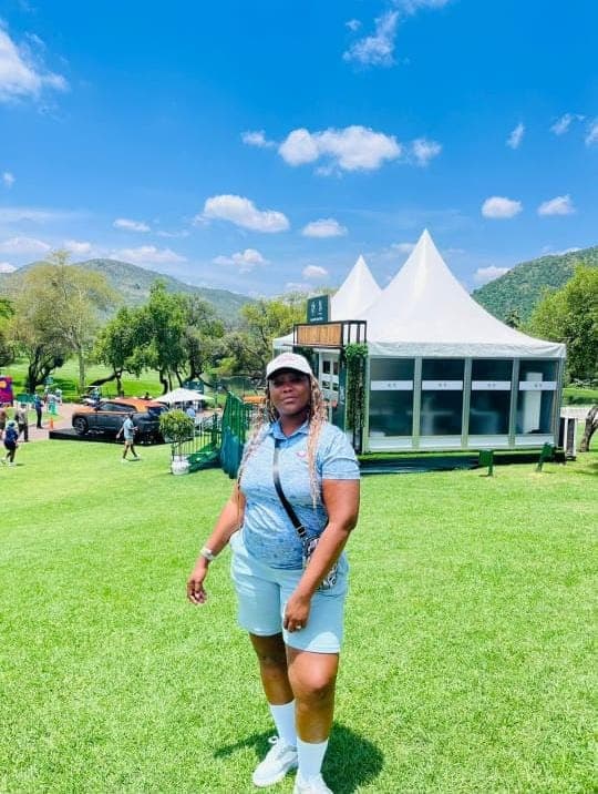 Woman in blue polo stands on a green lawn with white tents and mountains behind.
